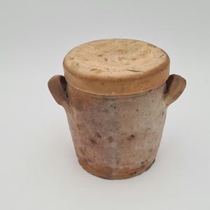 Op de afbeelding: Een kleine, rustieke, cilindrische keramische pot met een deksel. De pot is lichtbruin van kleur met een enigszins ruwe textuur en twee kleine handvatten. Het deksel is een lichtere bruintint. De pot is ongeveer 13 cm hoog.