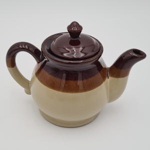 Może przedstawiać: Ceramiczny czajnik z pokrywką i uchwytem. Czajnik jest głównie beżowy z brązowymi i ciemnobrązowymi poziomymi paskami. Dzióbek i uchwyt są brązowe. Pokrywka ma mały, zaokrąglony guz.