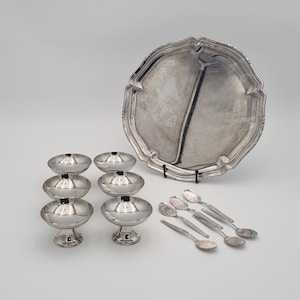 Puede incluir: Una colección de piezas de servicio de plata, que incluye una fuente grande y ornamentada, seis cucharas pequeñas y seis fuentes de varios niveles. Los artículos están dispuestos sobre una superficie blanca, reflejando la luz.
