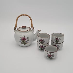 May include: A white ceramic tea set with a teapot and four teacups. The teapot has a woven handle and a brown lid. The set is decorated with a floral pattern in shades of purple, pink, and blue.
