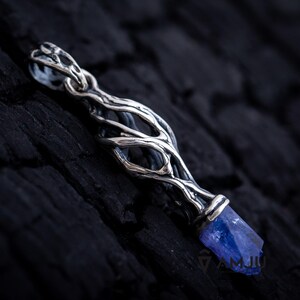 May include: A silver pendant with a blue gemstone set in a twisted, branch-like design. The pendant is on a dark, textured surface.