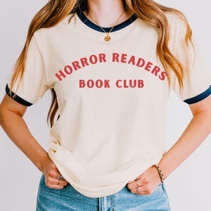 May include: Cream-colored t-shirt with navy blue trim and the words "HORROR READERS BOOK CLUB" in red. The shirt is paired with blue jeans. A gold necklace and bracelet are also visible.