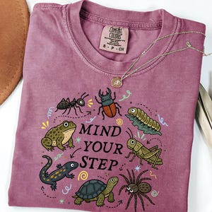 May include: A mauve short-sleeved t-shirt featuring a graphic print of various insects and small animals, including a toad, turtle, spider, and beetle, with the text "MIND YOUR STEP" in the center. The shirt has a crew neckline and a tag.