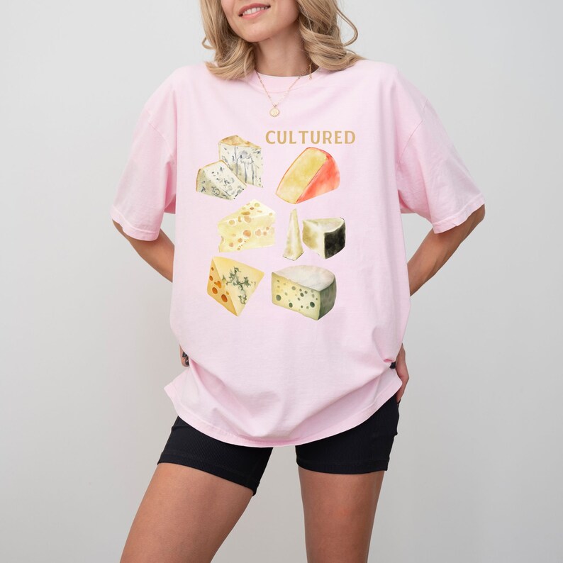 May include: A pink t-shirt with a graphic of different types of cheese and the word "CULTURED" in gold lettering.