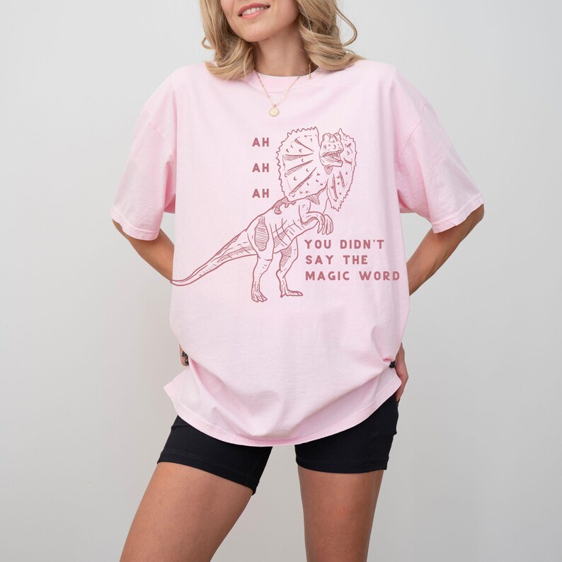 May include: A light pink t-shirt featuring a Dilophosaurus dinosaur illustration in a reddish-brown color. The text on the shirt reads "AH, AH, AH, YOU DIDN'T SAY THE MAGIC WORD."