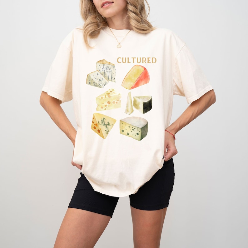 May include: A woman wearing a light beige t-shirt with a graphic of different types of cheese and the word "CULTURED" printed on it. She is wearing black biker shorts.