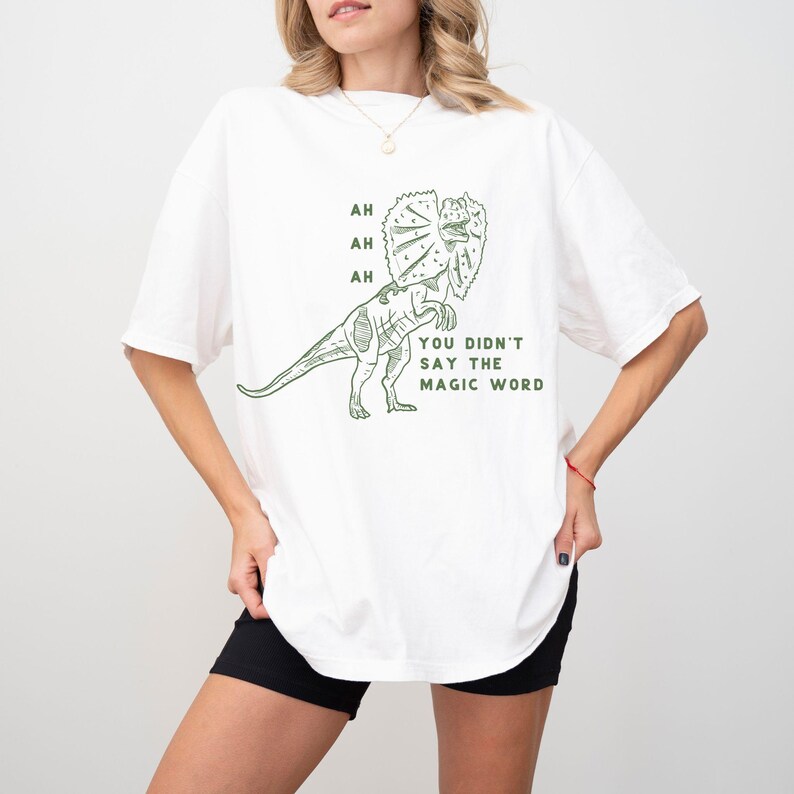 May include: White t-shirt with a green dinosaur graphic. The dinosaur has a frill around its neck and the text "AH AH AH" and "YOU DIDN'T SAY THE MAGIC WORD" in green. The shirt is a casual, oversized fit.