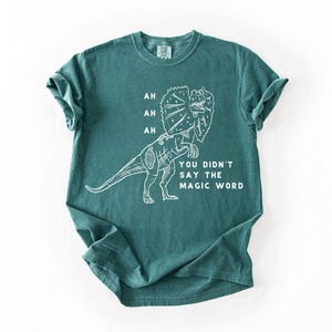 May include: Teal-colored t-shirt featuring a white line drawing of a Dilophosaurus dinosaur. The text on the shirt reads "AH, AH, AH, YOU DIDN'T SAY THE MAGIC WORD."
