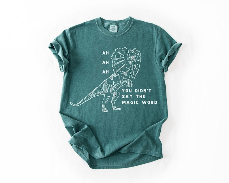May include: Teal-colored t-shirt featuring a white line drawing of a Dilophosaurus dinosaur. The text on the shirt reads "AH, AH, AH, YOU DIDN'T SAY THE MAGIC WORD."