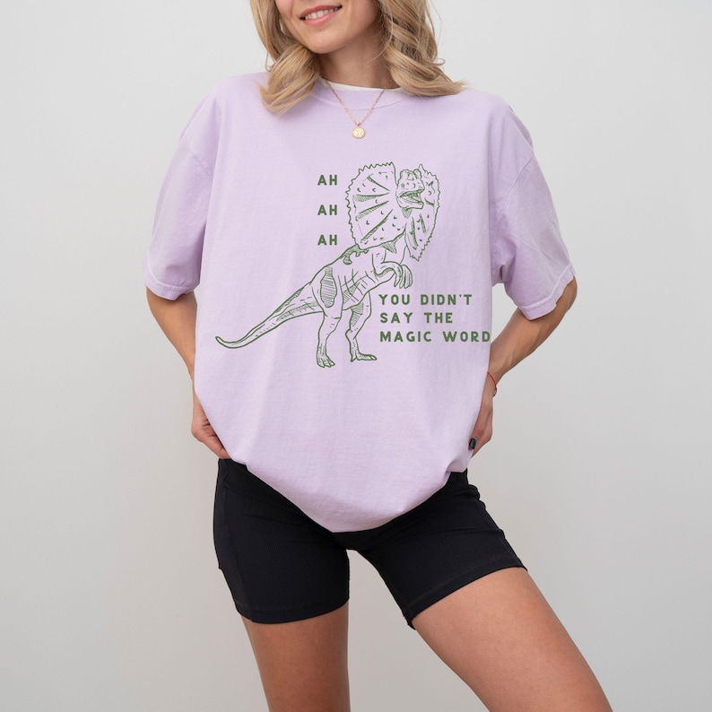 May include: A light purple t-shirt featuring a green dinosaur illustration with the text "AH, AH, AH, YOU DIDN'T SAY THE MAGIC WORD." The dinosaur has a frill around its neck. The shirt is a casual, oversized fit.