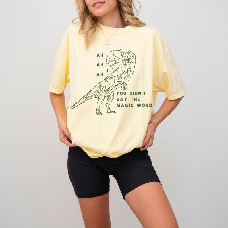 May include: A light yellow t-shirt with a green dinosaur graphic and the text "AH AH AH YOU DIDN'T SAY THE MAGIC WORD." The shirt has short sleeves and a relaxed fit. The model is wearing black shorts.