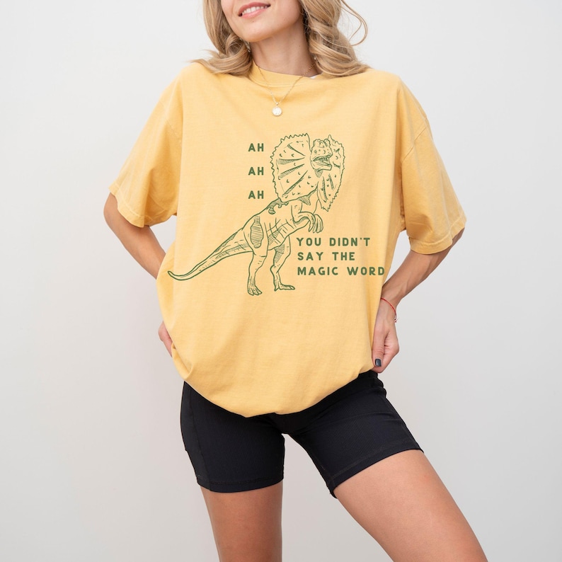May include: Mustard yellow t-shirt with a green dinosaur graphic. The dinosaur has a frill around its neck and the text "AH AH AH YOU DIDN'T SAY THE MAGIC WORD". The shirt is a relaxed fit.