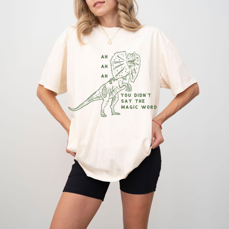 May include: Cream-colored t-shirt featuring a green Dilophosaurus dinosaur graphic with the text "AH, AH, AH, YOU DIDN'T SAY THE MAGIC WORD." The shirt has short sleeves and a relaxed fit.