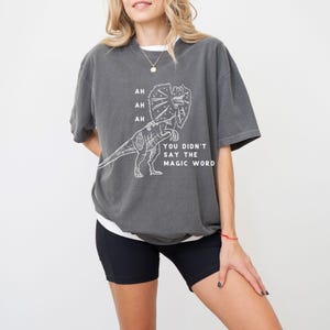 May include: Dark gray t-shirt with a white graphic of a Dilophosaurus dinosaur and the text "AH AH AH YOU DIDN'T SAY THE MAGIC WORD". The shirt is a relaxed fit and has short sleeves.