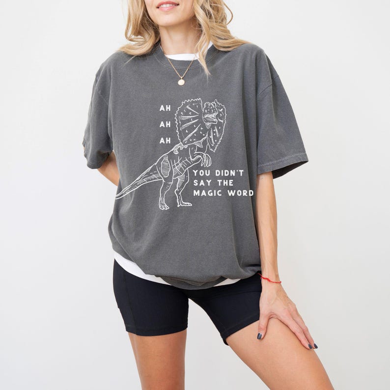 May include: Dark gray t-shirt with a white graphic of a Dilophosaurus dinosaur and the text "AH AH AH YOU DIDN'T SAY THE MAGIC WORD". The shirt is a relaxed fit and has short sleeves.