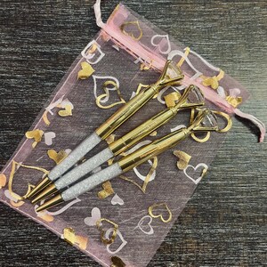 May include: Three gold glitter pens with diamond-shaped tops in a pink mesh bag with gold hearts printed on it.