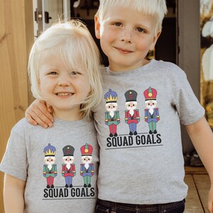 May include: Two children wearing gray t-shirts with a colorful nutcracker design and the text "SQUAD GOALS". The nutcrackers are in red, blue, green, and black uniforms. The children are smiling and standing close together.