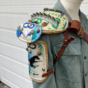 May include: Handmade shoulder and arm armor made from repurposed materials. The armor features license plates, metal spikes, and leather straps. The shoulder piece has the word "FASTEN" on it. The arm piece has a black dinosaur graphic.