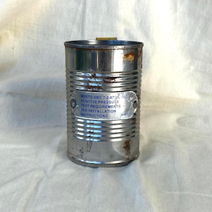 May include: A cylindrical metal canister with horizontal ridges and visible rust spots. A silver rectangular label is riveted to the front, featuring blue text that reads: MEETS UBC 7-2-97/UL 10C POSITIVE PRESSURE TEST REQUIREMENTS SEE INSTALLATION INSTRUCTIONS.