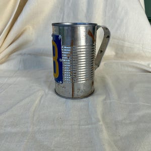 Op de afbeelding: Een cilindrische metalen mok gemaakt van een geribbelde conservenblik met een geklonken metalen handvat. Aan de zijkant is een blauw rechthoekig plaatje met een grote gele letter B bevestigd. Het metalen oppervlak vertoont roestvlekken.