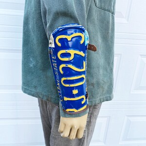 May include: A blue and yellow license plate arm guard with the text "STONE STATE 293" is shown on a mannequin arm. The arm guard is secured with a brown leather strap. The mannequin is wearing a green jacket and gray pants.