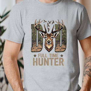 Puede incluir: Camiseta gris claro con un diseño de cabeza de ciervo y el texto "DAD FULL TIME HUNTER". La palabra "DAD" está en un patrón de camuflaje, y el texto es marrón. Prenda de vestir informal.