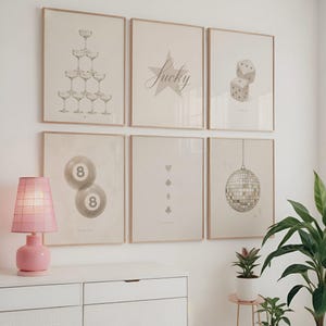 May include: A set of framed art prints in neutral tones. The prints depict champagne glasses, dice, a star with the word "Lucky", billiard balls, card suits, and a disco ball. A pink lamp is on a white chest of drawers.