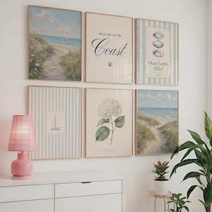 May include: A set of framed prints with coastal and botanical themes. The artwork includes beach scenes, a hydrangea illustration, and text reading "meet me on the Coast" and "Oyster Lovers Club". A pink lamp and potted plants are also visible.