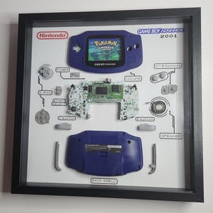 May include: Framed Game Boy Advance 2001 display. The display includes a disassembled blue Game Boy Advance, with components labeled and arranged on a white background. The game "Pokemon Emerald Version" is visible on the screen.