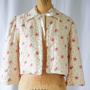 May include: Cream-coloured quilted jacket with a floral pattern of pink roses. The jacket has a collar, three-quarter sleeves, and a white ribbon tie closure. It is displayed on a vintage dress form.