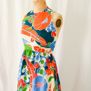 Pode incluir: Um vestido maxi sem mangas com uma estampa floral vibrante. O vestido apresenta um fundo branco com flores grandes de cor laranja, azul e verde. O vestido tem um corpete justo e uma saia fluida.