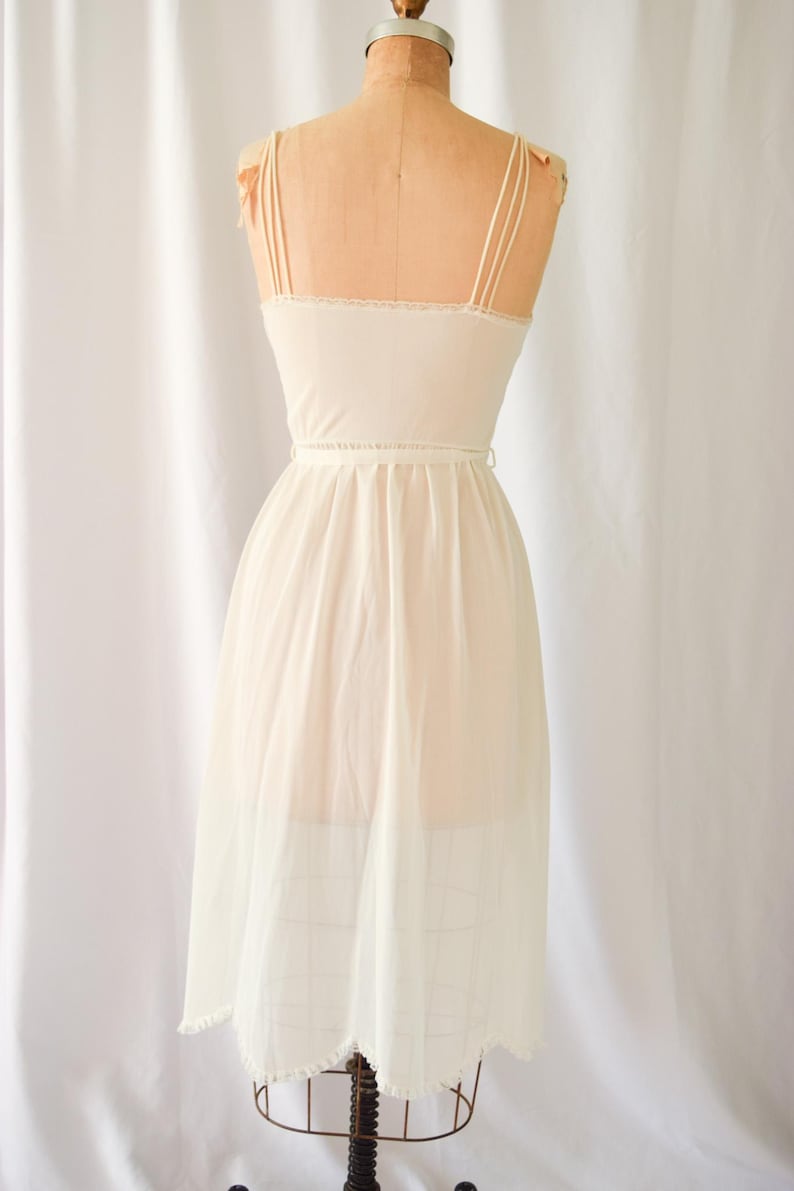 K&ouml;nnte beinhalten: Ein wei&szlig;es Schlupfkleid mit einem gewellten Saum und d&uuml;nnen Tr&auml;gern. Das Kleid hat ein tailliertes Oberteil und einen flie&szlig;enden Rock.