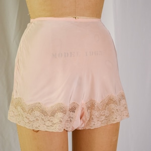 May include: Pink nylon shorts with a lace trim. The shorts have a label that reads "Model 1967".