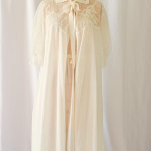 K&ouml;nnte beinhalten: Ein wei&szlig;es Spitzenkleid mit einer Schleife aus Satin. Das Kleid hat eine durchsichtige &Uuml;berlage und einen Spitzenbesatz.