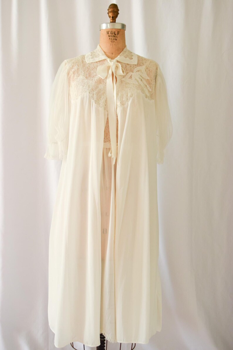 K&ouml;nnte beinhalten: Ein wei&szlig;es Spitzenkleid mit einer Schleife aus Satin. Das Kleid hat eine durchsichtige &Uuml;berlage und einen Spitzenbesatz.
