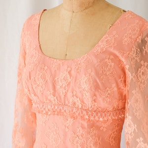 Puede incluir: Un vestido de encaje de color melocotón con mangas largas y un escote redondo. El vestido tiene un ribete de cuentas alrededor de la cintura.