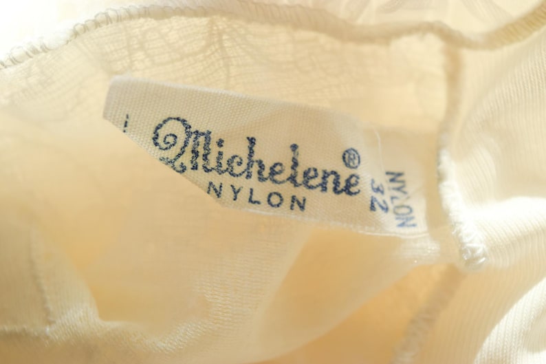 K&ouml;nnte beinhalten: Ein wei&szlig;es Nylon-Etikett mit dem Text "Michelene" und "Nylon 32" in blauer Schrift.