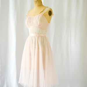 Op de afbeelding: Een lichtroze vintage jurk op een paspop. De jurk heeft een getailleerd lijfje met delicate bandjes en een zwierige rok. Witte kanten details sieren de taille. De jurk is gemaakt van een transparante stof.