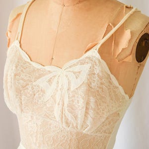 K&ouml;nnte beinhalten: Ein wei&szlig;es Spitzen-Schlafkleid mit Schleifen-Detail am Ausschnitt. Das Kleid hat verstellbare Tr&auml;ger und einen gerafften Rock.