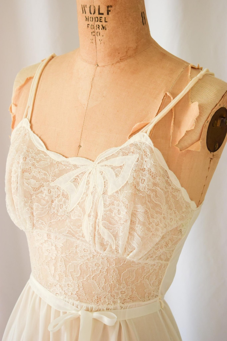 K&ouml;nnte beinhalten: Ein wei&szlig;es Spitzen-Schlafkleid mit Schleifen-Detail am Ausschnitt. Das Kleid hat verstellbare Tr&auml;ger und einen gerafften Rock.