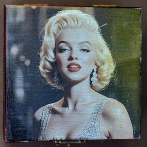 Può includere: Piccola scatola quadrata in legno con un'immagine vintage di Marilyn Monroe. L'immagine mostra Monroe con capelli biondi, rossetto rosso e orecchini scintillanti. La scatola ha un aspetto invecchiato e usurato.