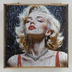 Op de afbeelding: Een close-up van een vrouw met blond haar die een rode jurk en een diamanten ketting draagt. De achtergrond is donkerblauw met zilveren glitter.