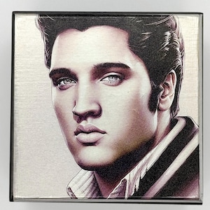 Könnte beinhalten: Quadratisches Kunstwerk mit einem Porträt von Elvis Presley. Das Bild zeigt eine Nahaufnahme seines Gesichts mit dunklem Haar und auffälligen blauen Augen. Er trägt eine gestreifte Jacke und ein Hemd mit Kragen. Das Kunstwerk ist in einem schwarzen Quadrat gerahmt.