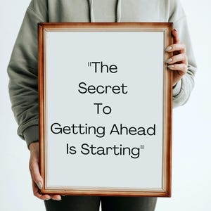 May include: A person holding a framed sign with the text "The Secret To Getting Ahead Is Starting".