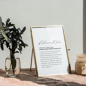 Pode incluir: Uma impressão emoldurada em ouro com o texto "Difference Maker" e uma definição da palavra. A impressão está sobre uma superfície rosa com um pequeno vaso de vegetação e uma vela.