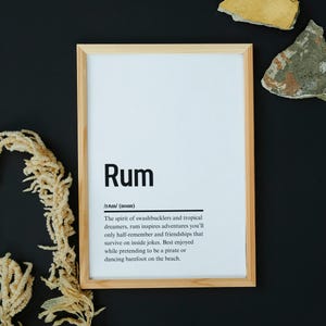 Op de afbeelding: Een ingelijst print met een witte achtergrond en zwarte tekst die het woord "Rum" definieert als de geest van swashbucklers en tropische dromers. De tekst beschrijft rum als inspiratie voor avonturen, vriendschappen en inside jokes. Het suggereert dat rum het best genoten wordt terwijl je doet alsof je een piraat bent of op het strand danst met blote voeten.