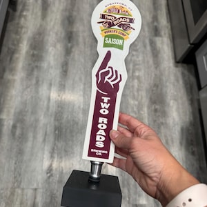 May include: A Two Roads Brewing Co. tap handle with a black base. The tap handle has a white top with the words "TWO ROADS" and "WORKER'S COMP SAISON". The handle features a burgundy hand pointing up.
