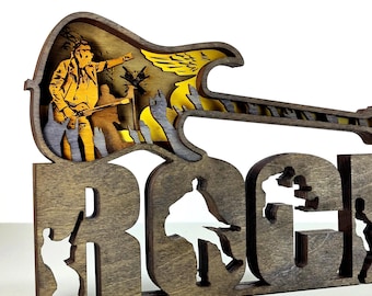 May include: Wooden sculpture featuring a guitar shape above the word "ROCK". The guitar design has a layered effect with a musician silhouette. The word "ROCK" has cut-out silhouettes of musicians. The sculpture is made of brown wood.