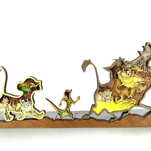 May include: A wooden Lion King-themed decorative piece. The artwork features Simba, Timon, and Pumbaa in a layered design. The characters are set against a backdrop of a savanna landscape, with yellow and brown tones.