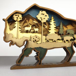 Puede incluir: Una escultura de bisonte de madera multicapa que representa una escena forestal. La escultura presenta árboles, ciervos, osos y montañas en tonos de marrón, verde y azul. El bisonte está de pie, con un diseño recortado detallado.